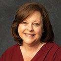 Sheila is pictured from the shoulders up wearing a garnet shirt. She has pale skin, short brown hair and is smiling at the camera. 