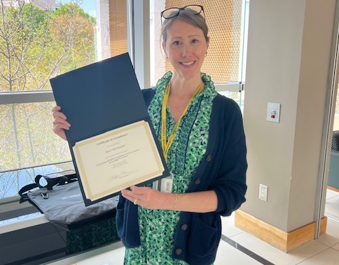CTE Certificate Completer Stacy Winchester Woman standing in hallway with windows holding a certificate.