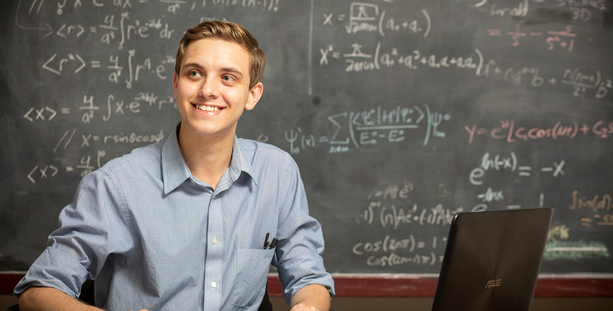 A student sits next to an open laptop in front of a blackboard full of complex mathematical equations.