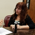 Andrea L'Hommedieu, of the Department of Oral History, sits at a table while interviewing a subject. The subject is mostly out of frame, but four fingers are visible on the left side of the image. A small recording device and some papers are on the table between them.