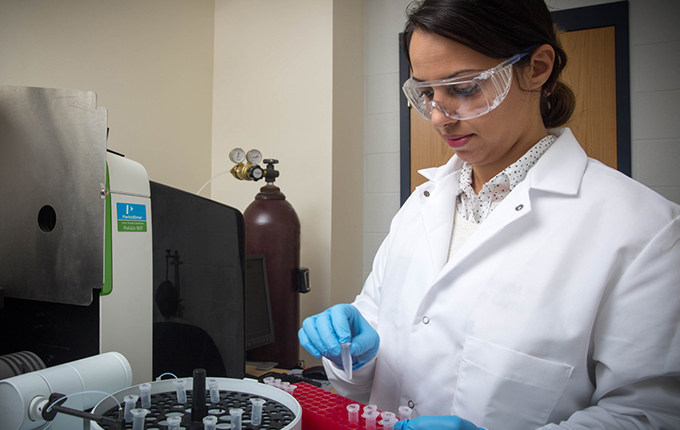 Female student in white lab coat, blue latex gloves, and safety glasses works with a tray of lab vials