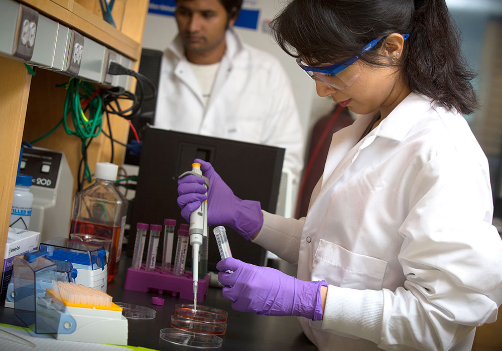 Graduate student pipetting into a tube wearing lab coat, safety goggles and gloves. 