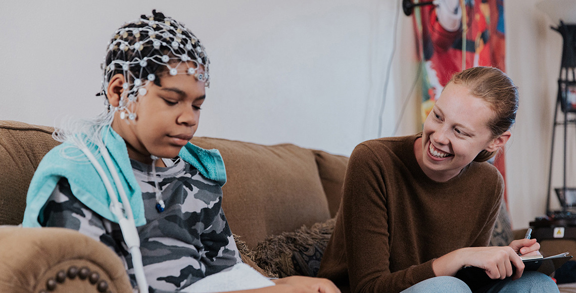 Wearing an EEG net on his head, AJ Peterman sits on his couch beside research assistant Ashlan Cheever, who is engaging him in conversation.