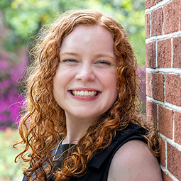 Headshot of a red-haired graduate smiling next to a brick wall and blooming flowers.