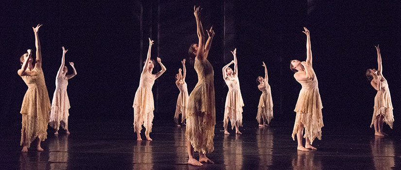 Dancers performing on stage, dressed in cream-colored long dresses, reaching up.