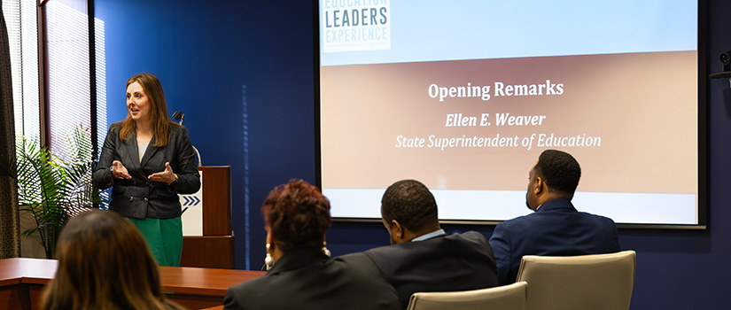 A woman in a green skirt adresses a room of people. A screen to the right reads "Opening Remarks: Ellen E. Weaver, State Superintendent of Education"