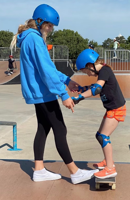 Chase Barclay helps a young girl balance on a skateboard at a skate park, both wearing helmets and protective gear.