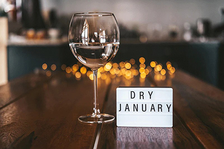 Wine glass on a wooden table next to a lightbox sign reading ‘Dry January,’ with soft, warm lights blurred in the background.