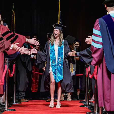 A University of South Carolina graduate wearing a cap and gown with a blue ZTA stole walks down a red carpet, smiling and high-fiving faculty members lined on both sides during the College of Hospitality, Retail and Sport Management Degrees of Excellence ceremony.