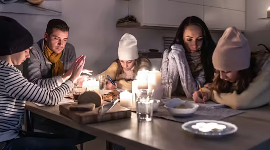 A family of 5 bundled up in knit scarves and hats gathers around a candle-lit dining table.