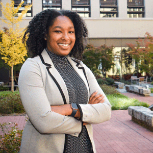 Glynnis Hagins wearing a black dress and cream and cream jacket stands in a brick courtyard with stone benches and fall trees in the background.