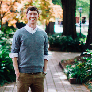 A man in a gray sweater stands on the brick walkway of the horseshoe with trees in the background