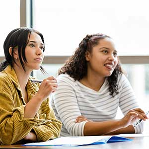 Two female students sit in a classroom with large windows.