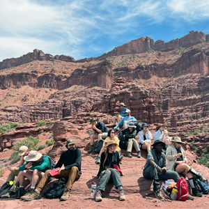 a group of a dozen people sit at the foot of large red rock formation 