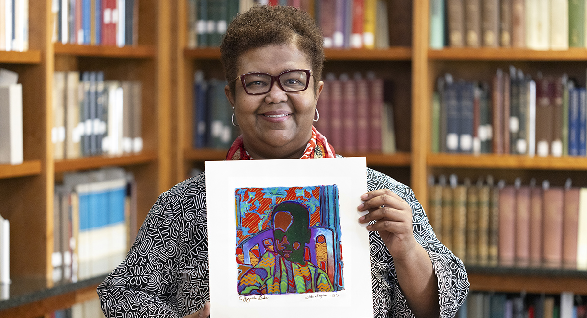 Nicole Cooke holds a 12x12 scrapbook page with a colorful, abstract drawing by John Steptoe, depicting what appears to be an African American man or child. 